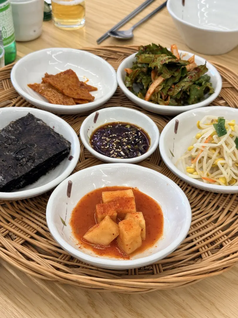 Mugeunji galbi-jjigae stew in white bowl surrounded by side dishes on woven table mat