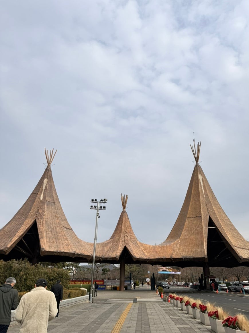 Three large teepee-style wooden roof structures forming the main entrance gate of Let's Run Park Seoul, with visitors walking toward the entry plaza and parking lots visible to the right