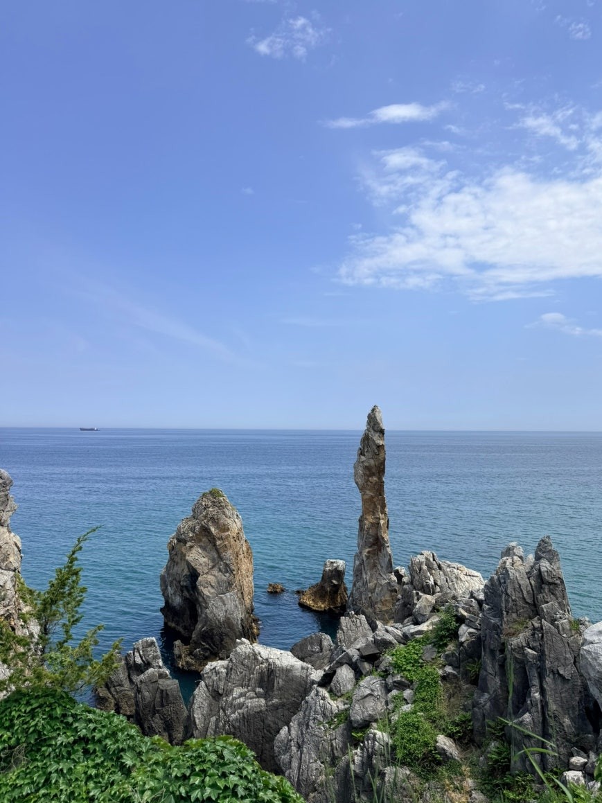 Chuam Candlestick Rock spire rising from jagged grey coastal rocks and green coastal plants, the East Sea stretching behind it under a clear sky