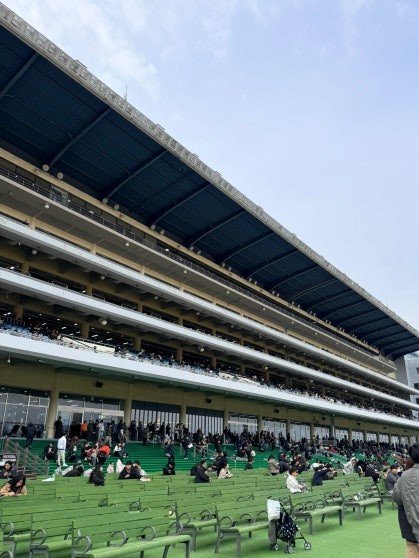 Multi-tiered concrete grandstand at Let's Run Park Seoul viewed from outside, with green painted seating tiers and outdoor benches occupied by visitors on a clear day