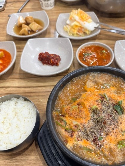 Abai-style blood-sausage soup (아바이순대국) served in a black stone hot pot bubbling with rich reddish-orange broth, with a small bowl of steamed white rice, red pepper paste, kimchi, and side dishes on a wooden table