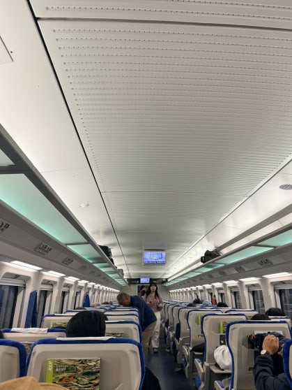 KTX-Eum train interior looking toward the front: rows of blue-and-white reclining seats, overhead luggage racks, and passengers in a well-lit carriage