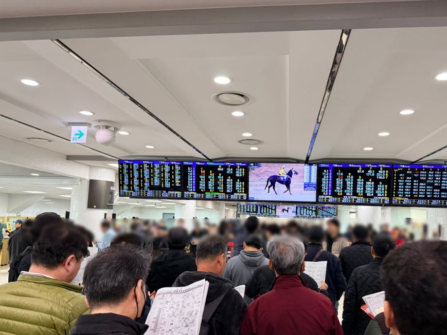Crowded second floor interior at Let's Run Park Seoul with multiple large digital odds boards spanning the ceiling, visitors standing and holding race programs, and a center screen showing a racing horse