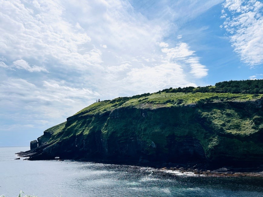 Udo Peak (우도봉) — steep green volcanic cliff with a small white lighthouse at the summit, rocky shoreline and calm blue sea in the foreground