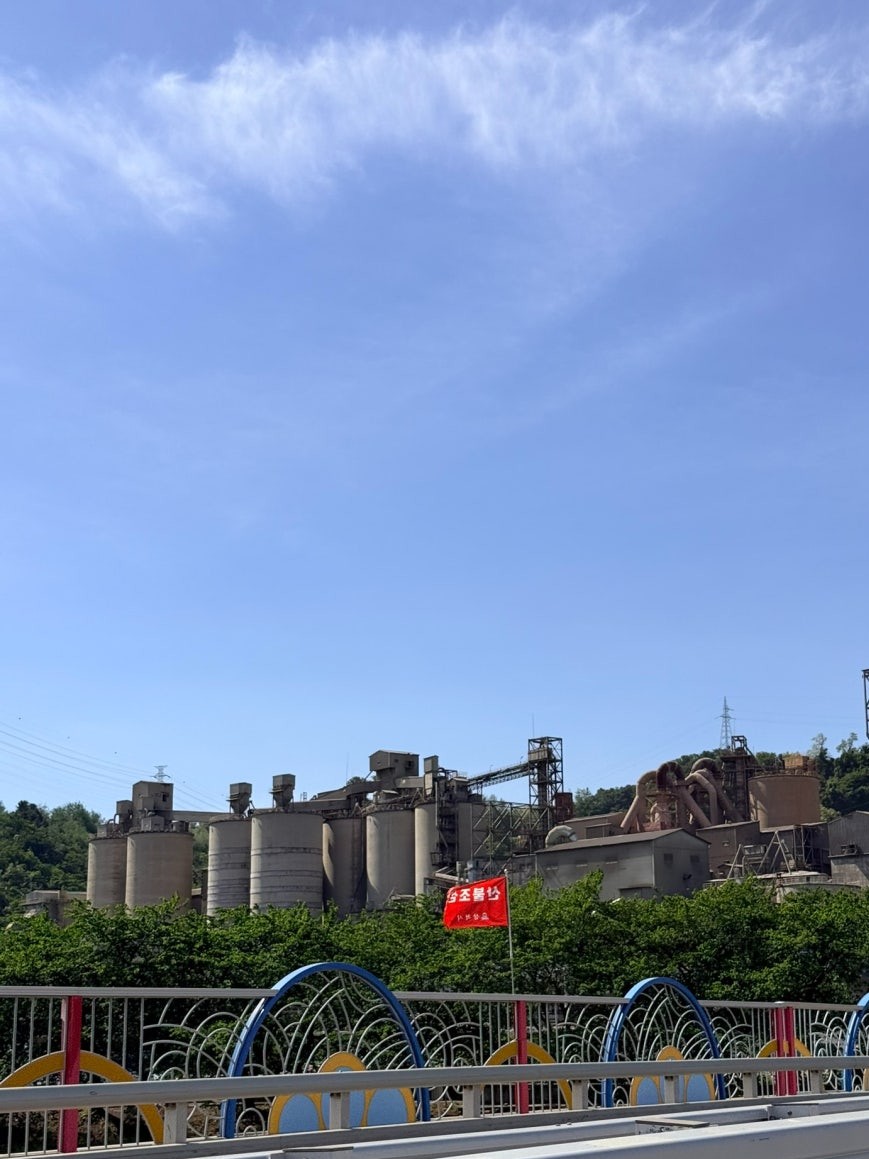 Cement factory with tall silos and metal industrial scaffolding towering above a green tree line, photographed from a colorfully railed bridge near Samcheok Station under a clear blue sky