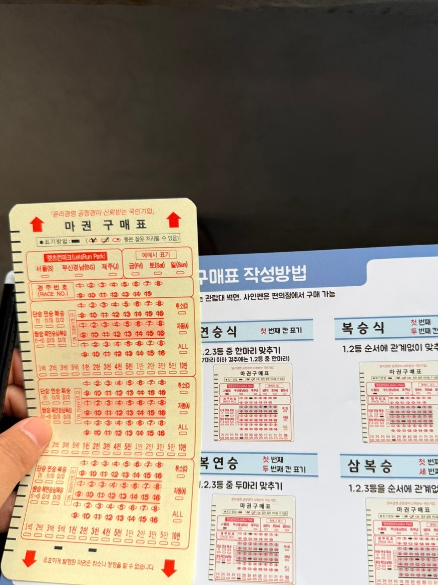 OMR betting card for Let's Run Park Seoul held in hand showing bubble-fill sections for race and horse numbers, with an instructional poster behind showing betting type categories