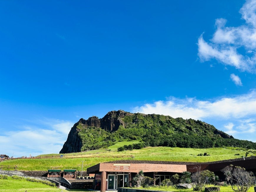 Seongsan Ilchulbong (성산일출봉) volcanic crater with a jagged rocky peak rising above a green hillside under deep blue sky, low visitor center building with grass courtyard in the foreground