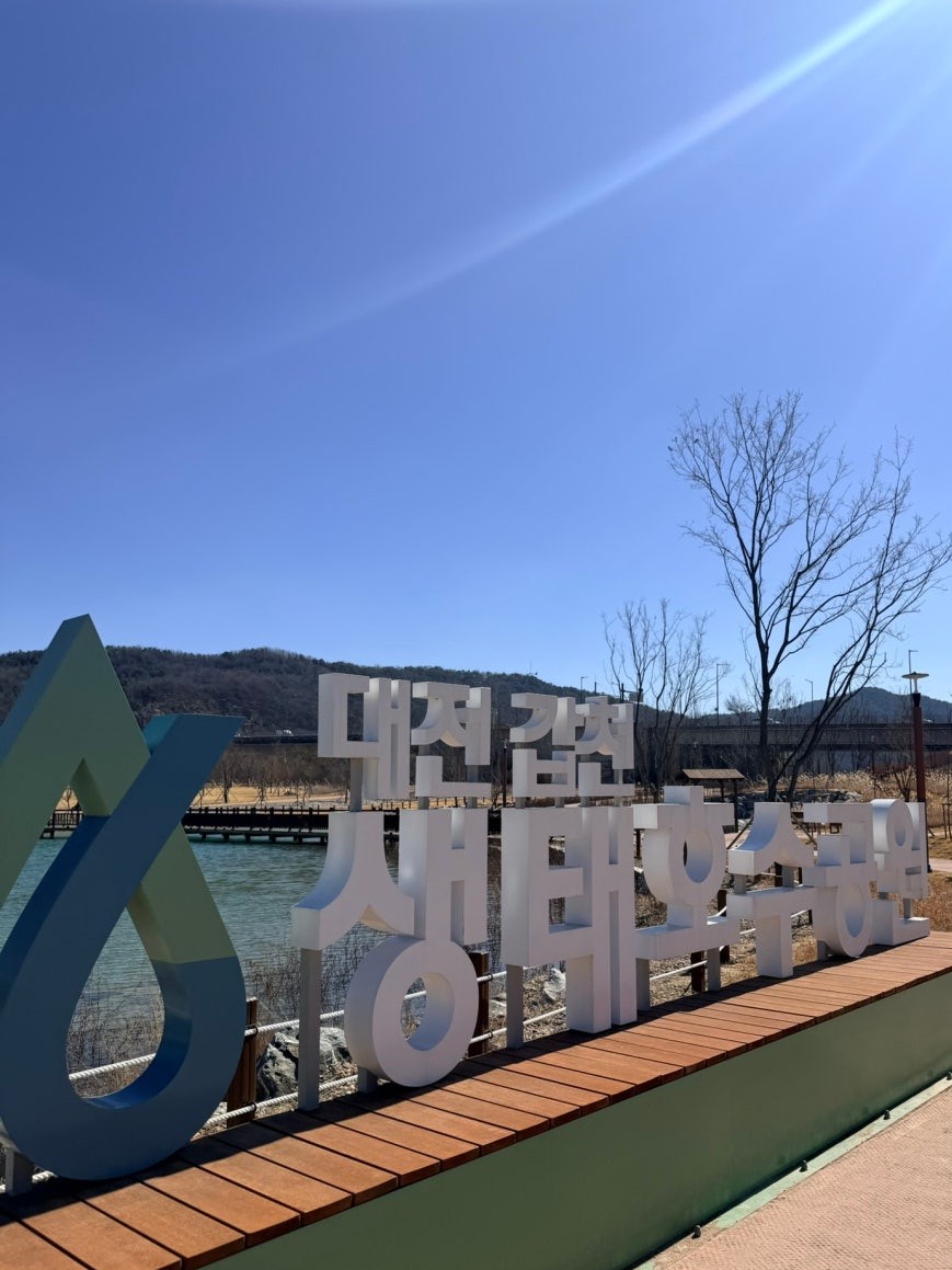Large white outdoor sign reading 대전 갑천 생태호수공원 at the park entrance with a blue logo to the left, the calm lake and a wooden pier visible behind under a clear blue winter sky
