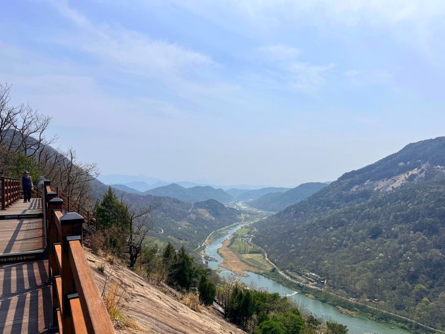 Elevated wooden deck walkway built along the sheer granite face of Yonggwolsan, with a panoramic view of the Seomjin River winding through forested mountain ridges below, and one hiker visible at the far end of the railing