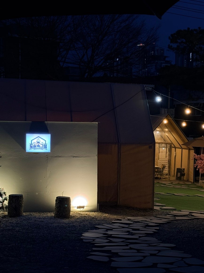 Nighttime exterior of Dosim Sok Grill in Siheung with an illuminated blue logo sign on a wall, string lights, and warm-lit glamping tent structures visible through the gate passage