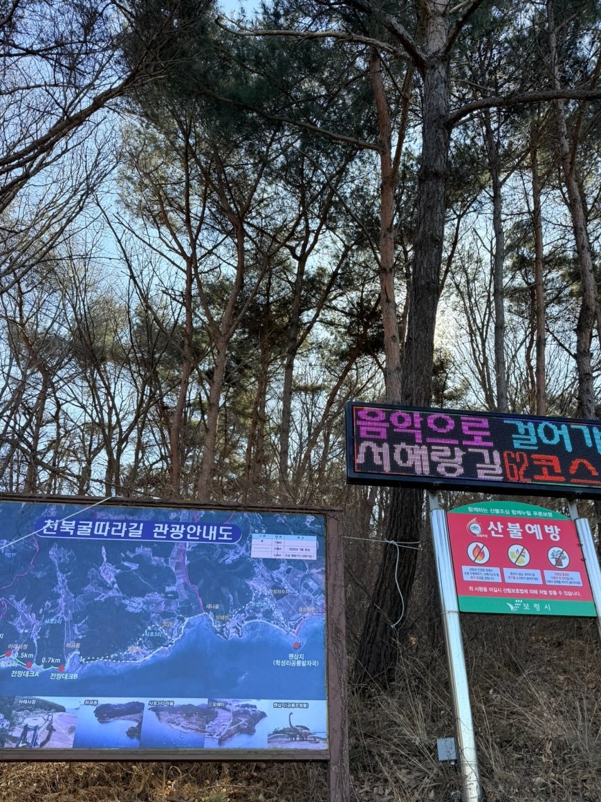 Trail entrance signs for Cheonbuk Gulttara-gil beside bare winter trees: a tourist map board showing the coastal walking route and an LED scrolling display reading 서해랑길 62코스 (Seohaerang-gil Trail Course 62)