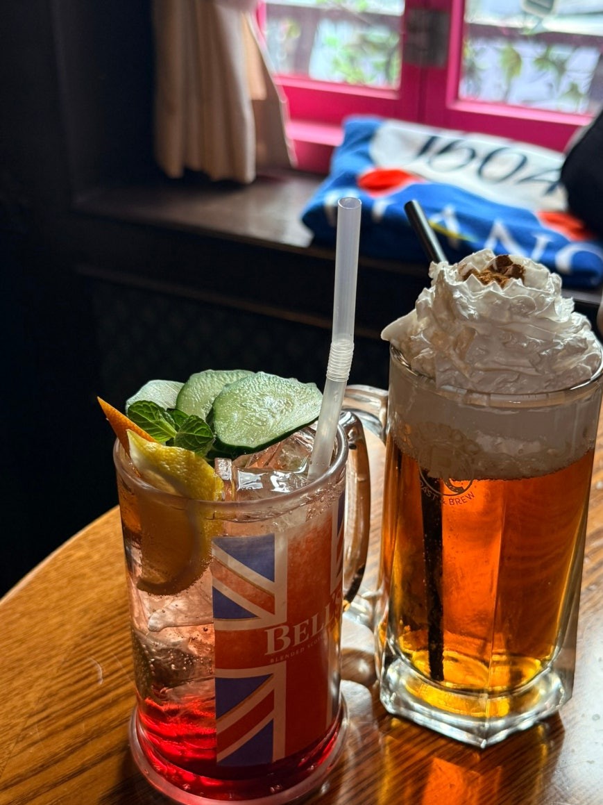 British gin highball in a Union Jack-branded glass with cucumber, mint, and citrus garnish, alongside butter beer in a large mug topped with whipped cream, on a wooden table