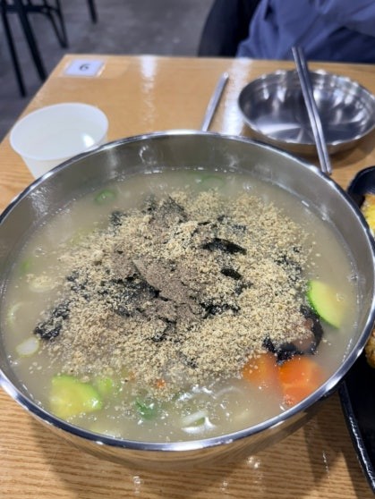 Bowl of gamja-ongsimi potato dumpling soup in a large silver metal pot, generously topped with ground perilla seeds and torn dried seaweed, with carrot and zucchini pieces visible in the milky white broth