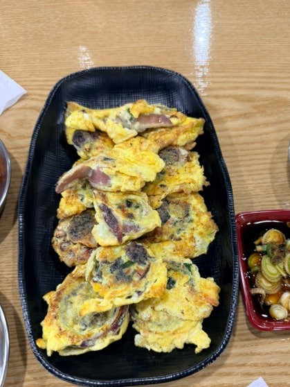 Plate of egg-battered squid sausage pieces arranged on a black rectangular serving tray, with green onion and filling visible inside the batter, accompanied by a small dish of scallion kimchi