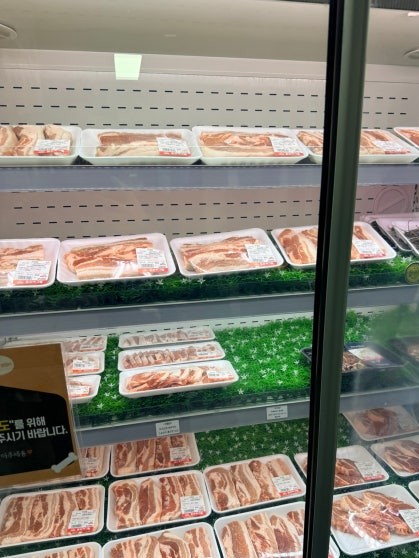 Refrigerated glass display case at Dosim Sok Grill showing multiple trays of raw pork belly cuts arranged on green garnish on the shelves with Korean price labels