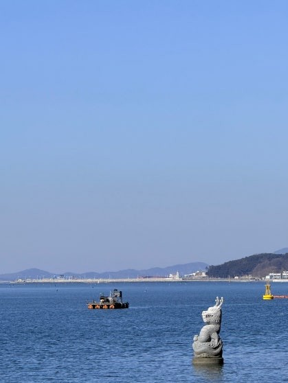 View from the coastal deck viewpoint at Cheonbuk showing a stone sea-creature sculpture standing in calm blue-green water, an orange fishing boat in the background, with distant mountains and harbor visible across the bay