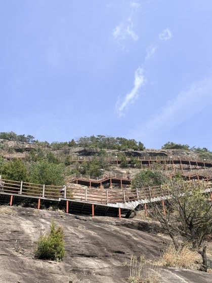 Yonggwolsan Sky Trail wooden deck structure viewed from the base of the cliff, showing multiple tiers of brown railings built into the bare granite cliff face under blue sky
