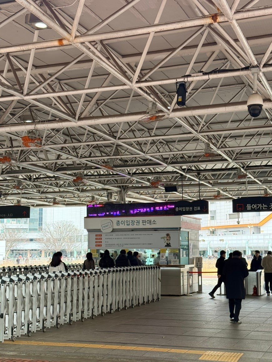 Paper ticket booth at Let's Run Park Seoul entrance, with folding metal turnstile barriers, visitors queuing at the counter, and a bilingual entrance sign