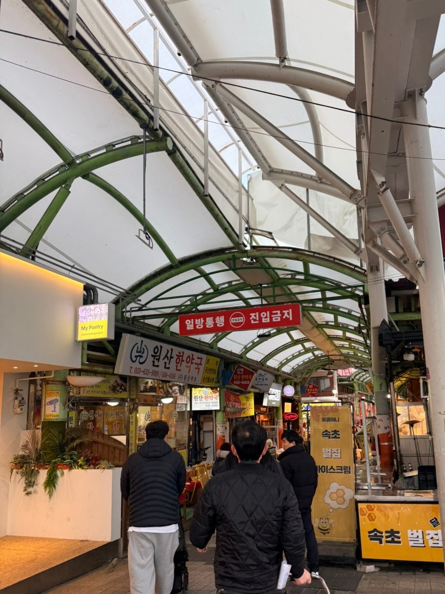 Covered arcade entrance to Sokcho Jungang Market with a red one-way traffic sign overhead, a Korean traditional medicine pharmacy and honey vendor stalls on either side, three visitors walking into the inner market corridor beneath a green steel arch roof