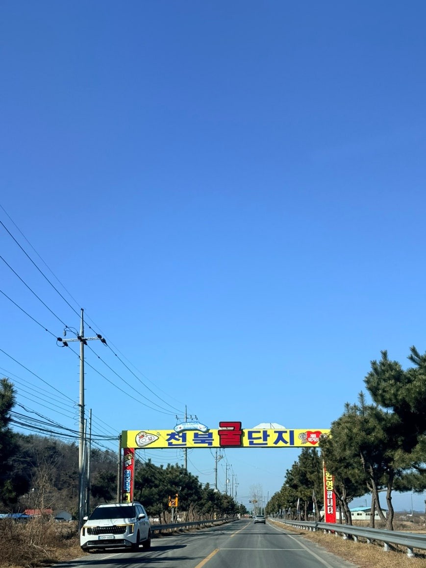 Road approach to Cheonbuk Oyster Village under a clear blue winter sky, with a large yellow and red 천북굴단지 welcome arch spanning the road and pine trees lining the right side