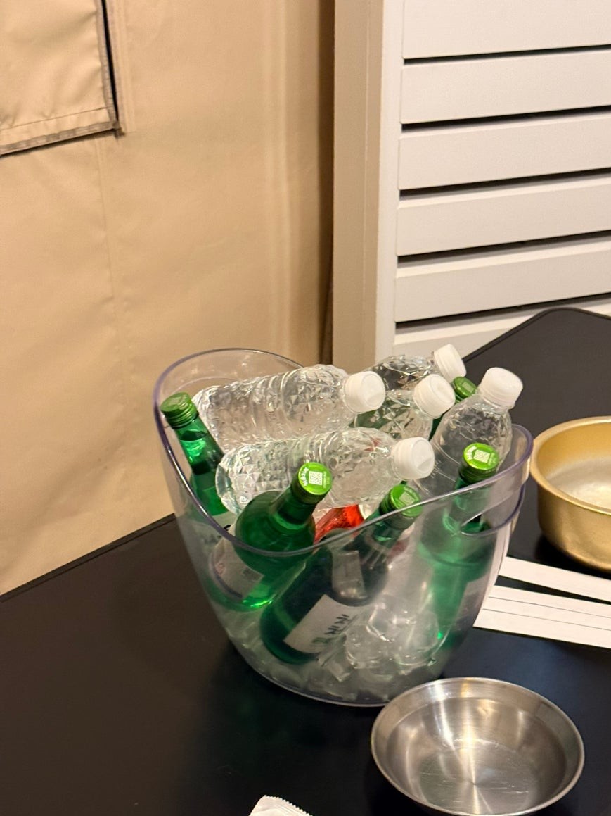 Clear plastic ice bucket filled with green soju bottles and clear water bottles packed in ice on a dark side table inside the tent, with Korean metal bowls nearby