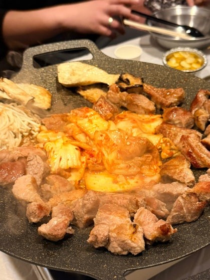 Round grill plate with pork belly pieces arranged around sizzling kimchi in the center, with enoki mushrooms and other items on the outer edge, sauce bowls visible in the background