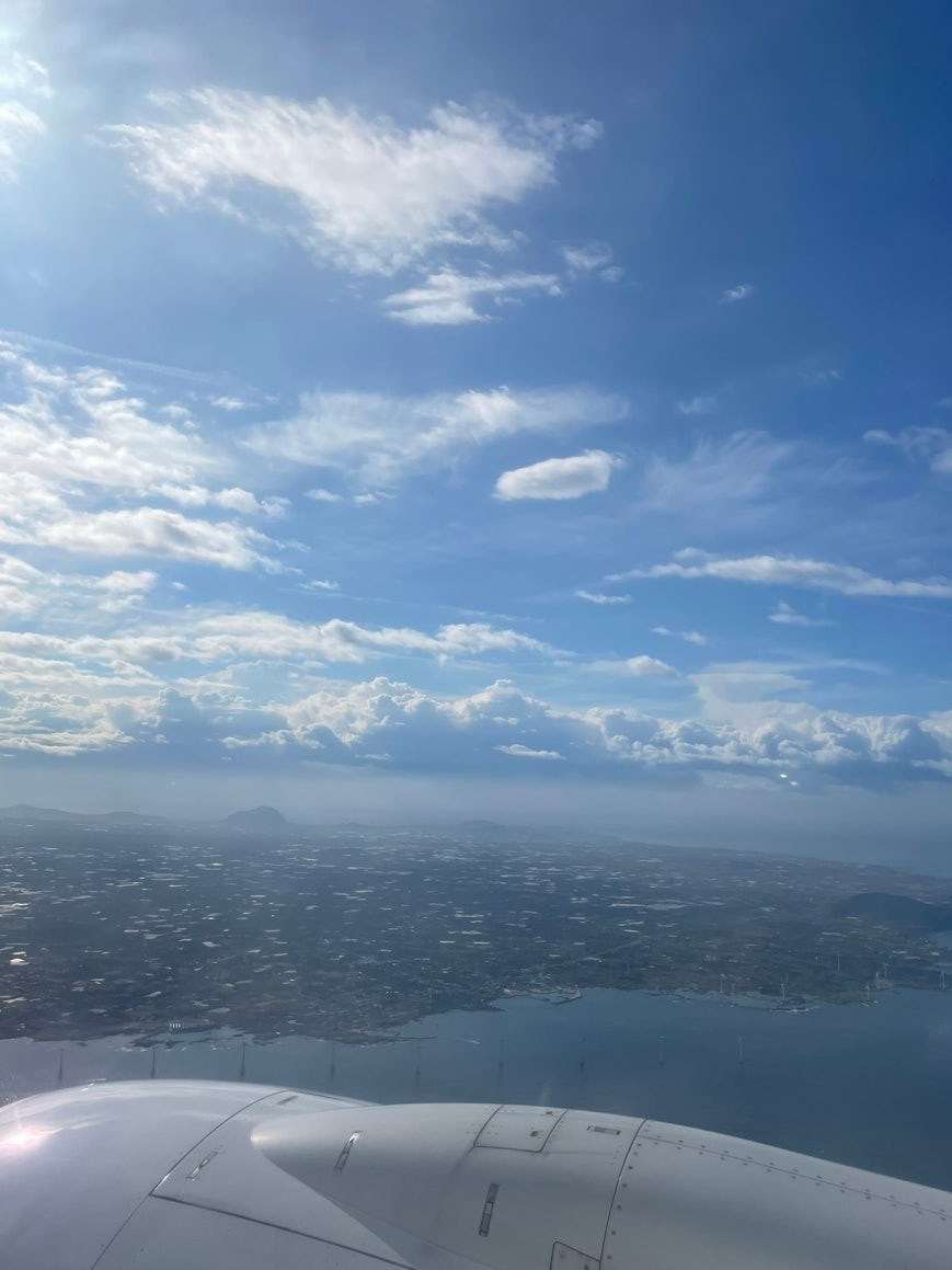 Aerial view from a plane window showing Jeju Island's coastline and city spread below, the engine nacelle in the foreground, and a faint peak visible on the horizon under a partly cloudy blue sky