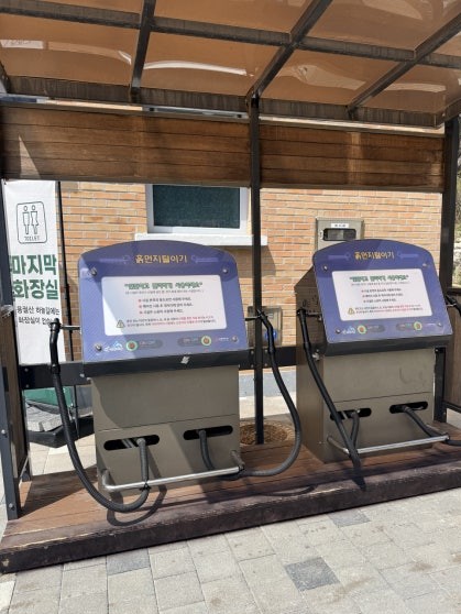 Two automated dust-removal machines installed side by side under a small open canopy near the Yonggwolsan trail entrance, with Korean signage reading 흙먼지 털이기 on the brick wall behind them and a last-restroom notice sign to the left