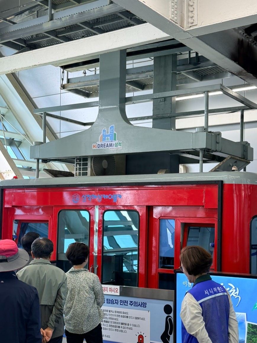 Red Samcheok Marine Cable Car cabin at the indoor boarding platform with passengers waiting at the door and a staff member in a blue vest on the right