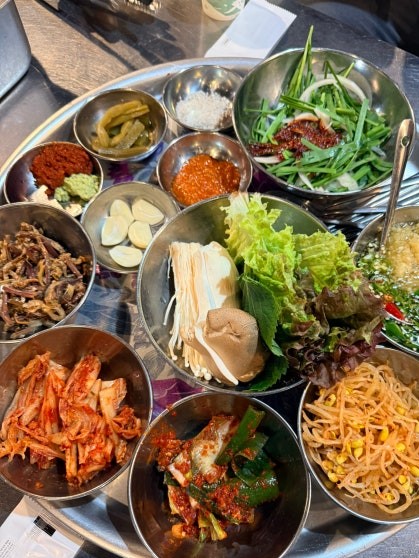 Round platter holding an assortment of banchan (반찬, Korean side dishes) in stainless steel bowls — kimchi, seasoned greens, enoki mushrooms, lettuce leaves, sliced ingredients, and a scallion salad arranged around a center plate with vegetables