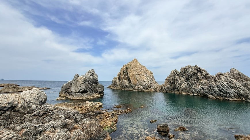 Three jagged rock formations rising from a shallow turquoise cove at Janghohang, with rough coastal rocks in the foreground under a partly cloudy sky