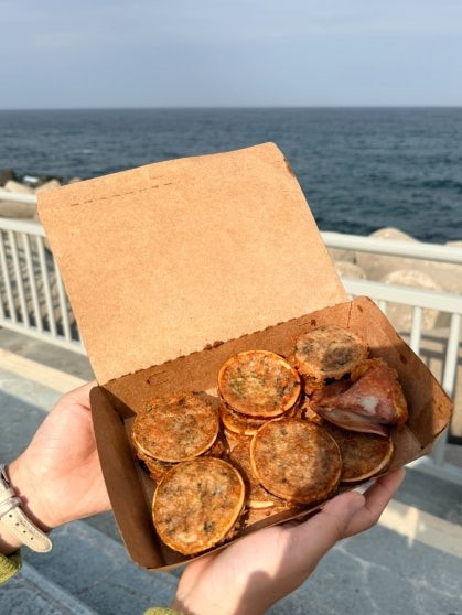 Open kraft paper takeout box of ojingeo sundae (오징어순대) held over a railing with the East Sea behind — sliced rounds of stuffed squid with one whole tapered tip in the box