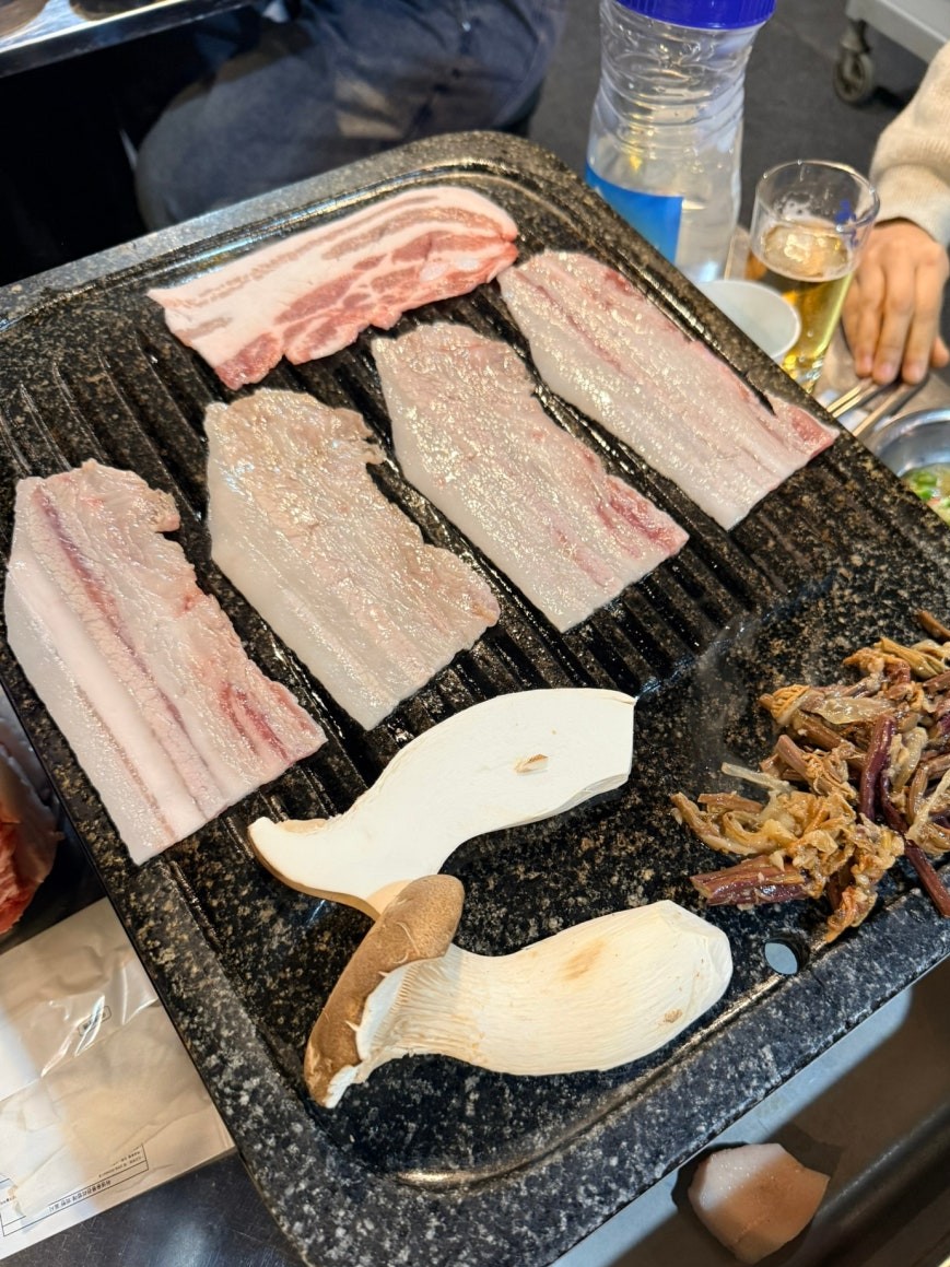 Grill plate with raw saeng-daepae pork belly slices in various cuts alongside king oyster mushrooms and a serving of braised namul (나물) on the right side of the grill, with side dish bowls at the edge of the frame