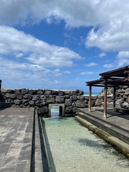 Gwamul Noecheontang outdoor freshwater spring bath enclosed by volcanic stone walls, with a shallow rectangular pool of clear water, a wooden pergola overhead, and the sea visible through a narrow gap in the far wall