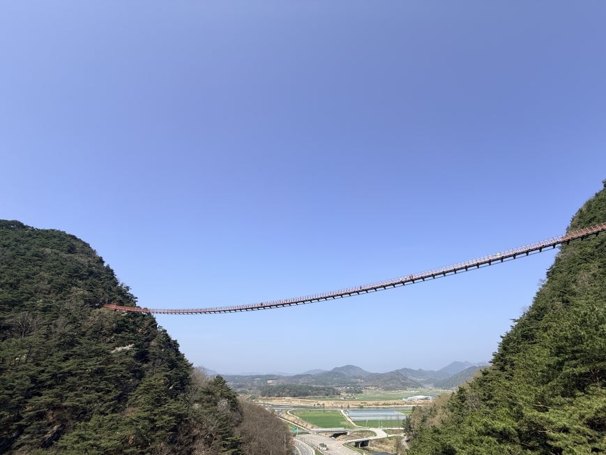 Chaegyesan Suspension Bridge seen from the midpoint observation deck, a thin red span connecting two forested mountain ridges, with a rural valley, road, and farmland below under clear blue sky