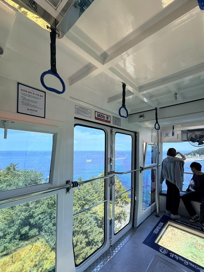 Interior of the Samcheok Marine Cable Car on the return journey, two passengers at panoramic windows with the deep blue East Sea and a forested coastal headland visible below
