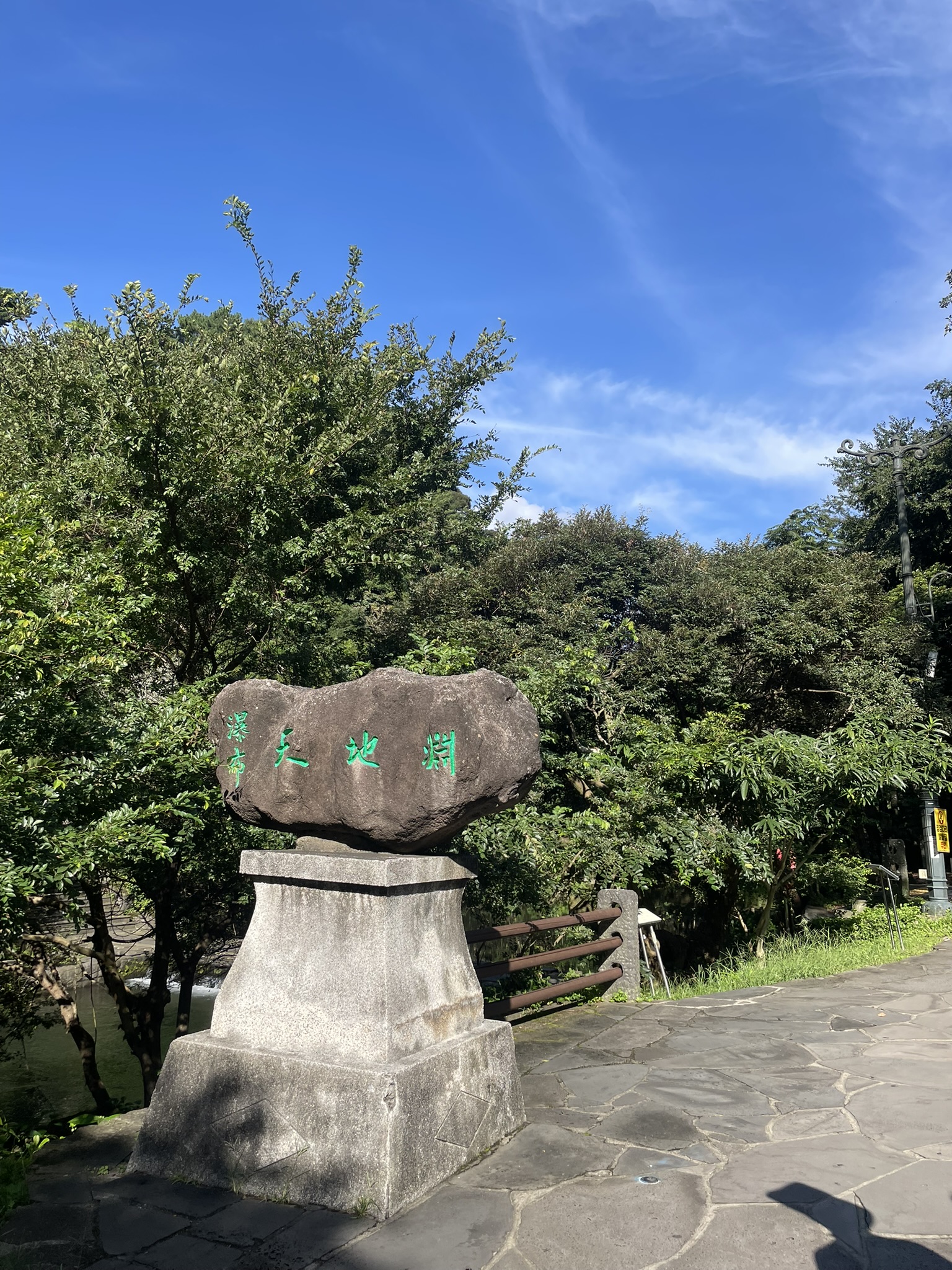 Stone entrance marker for Cheonjiyeon Falls (천지연폭포) with Korean characters in green carved on a dark volcanic boulder set on a concrete pedestal, blue sky and lush green trees visible behind