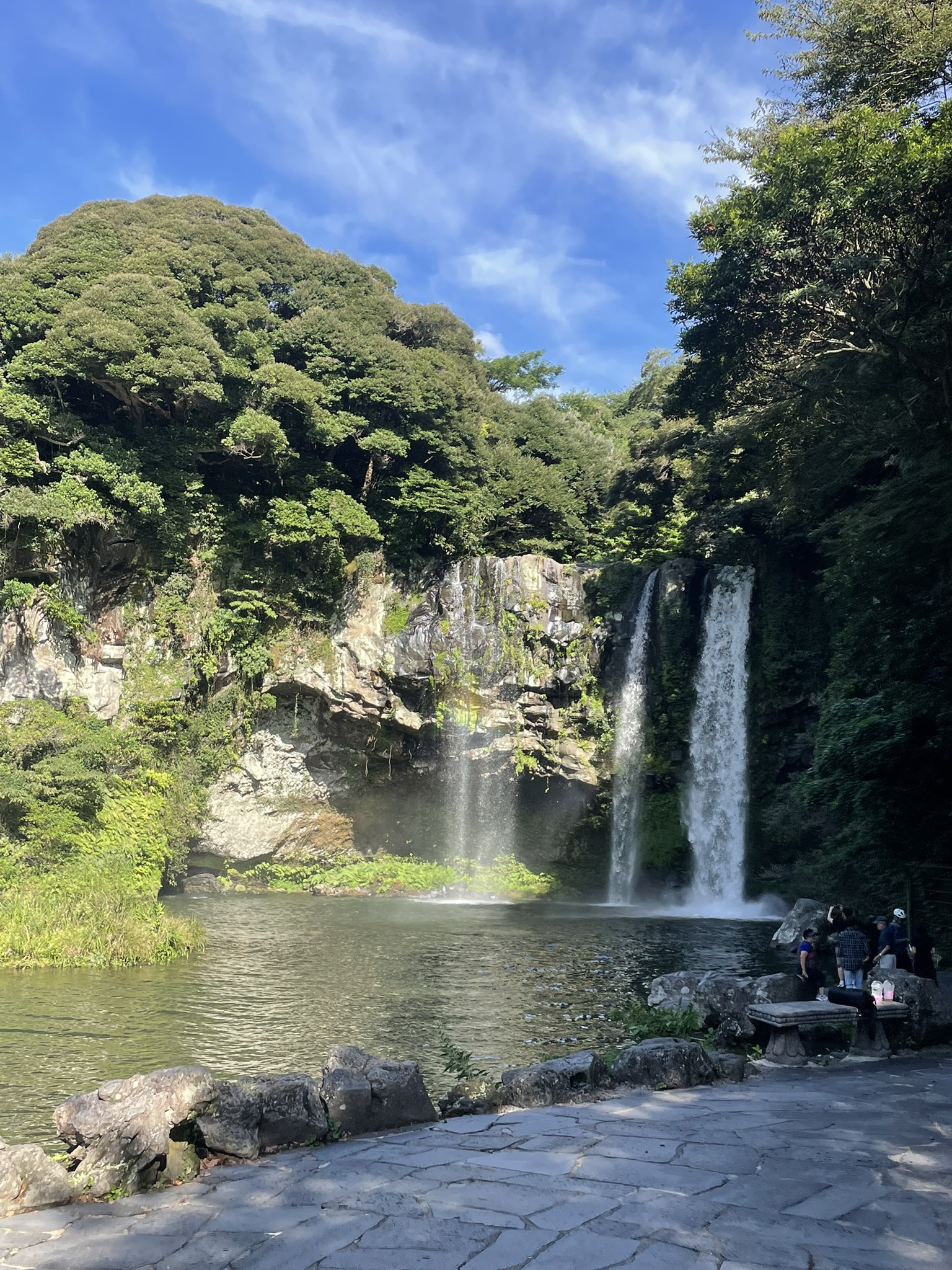 Cheonjiyeon waterfall (천지연폭포) cascading down a tall basalt and vegetation-covered cliff face into a wide green rock pool, stone-paved viewing terrace in the foreground and visitors at the rocks to the right