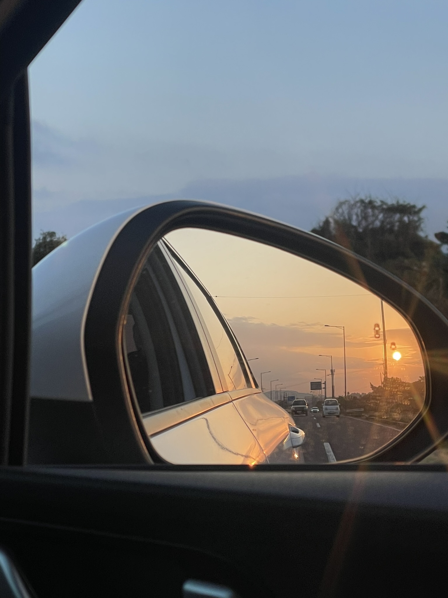 Early morning orange sunrise reflected in a silver car side mirror, a multi-lane urban road with vehicles and streetlights visible in the warm reflection against a grey pre-dawn sky