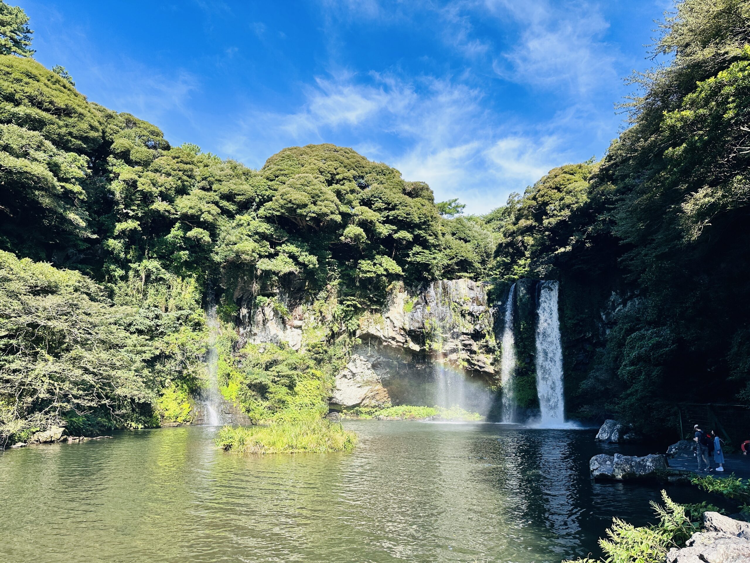 Two waterfalls — one wide curtain on the right, one narrower plume on the left — cascading into a broad emerald natural pool at Cheonjiyeon (천지연폭포), Seogwipo, a small forested island at the pool's center, dense green trees framing the scene under blue sky