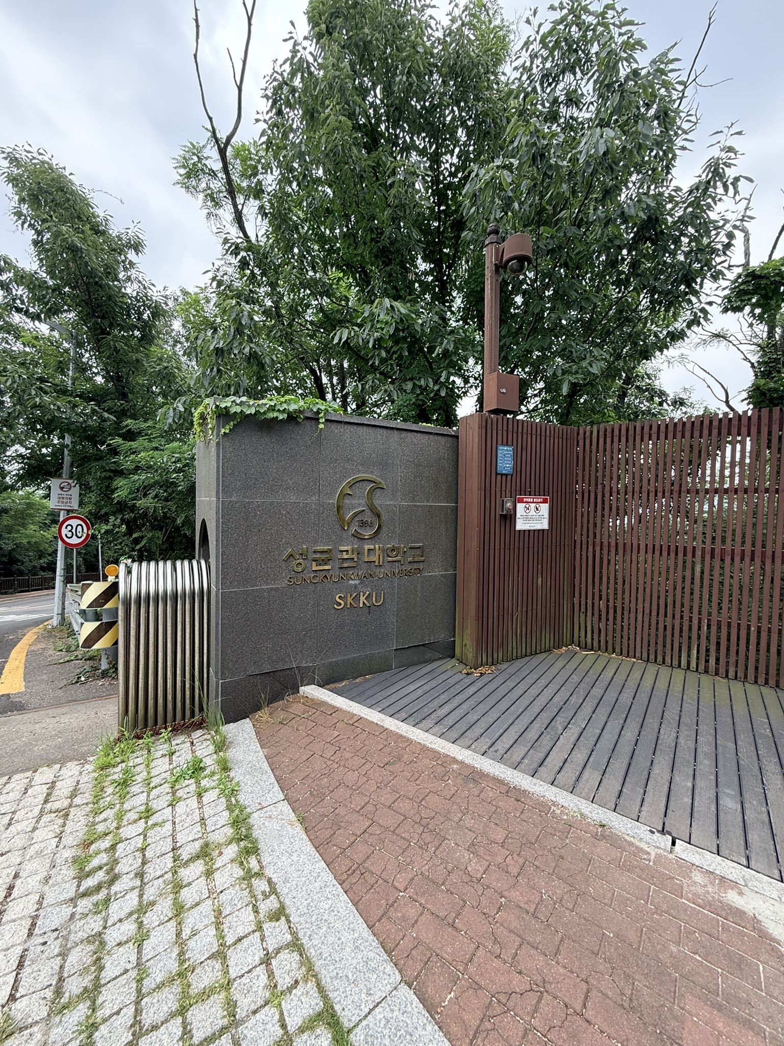 SKKU Sungkyunkwan University side gate with dark granite monolith bearing gold university crest and brown wooden fence on the approach road to the Bugaksan Malbawi trailhead