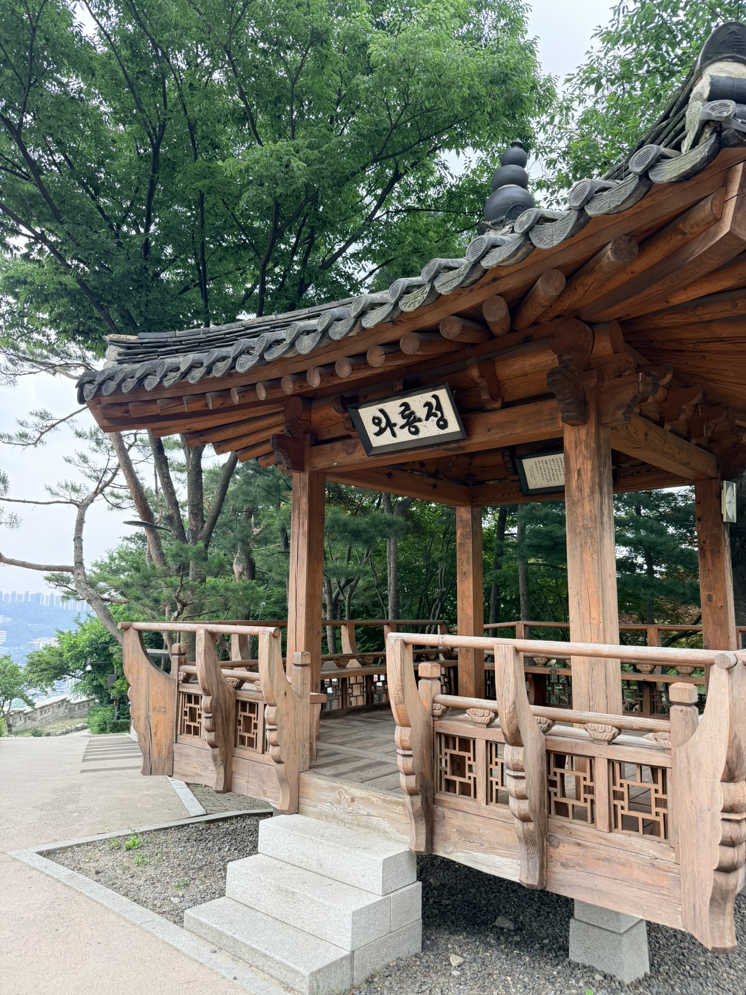 Traditional Korean open-air Waryong Pavilion with gray clay tile roof, carved geometric lattice wooden railings, stone steps, surrounded by dense summer trees, with a distant city skyline glimpsed through the foliage