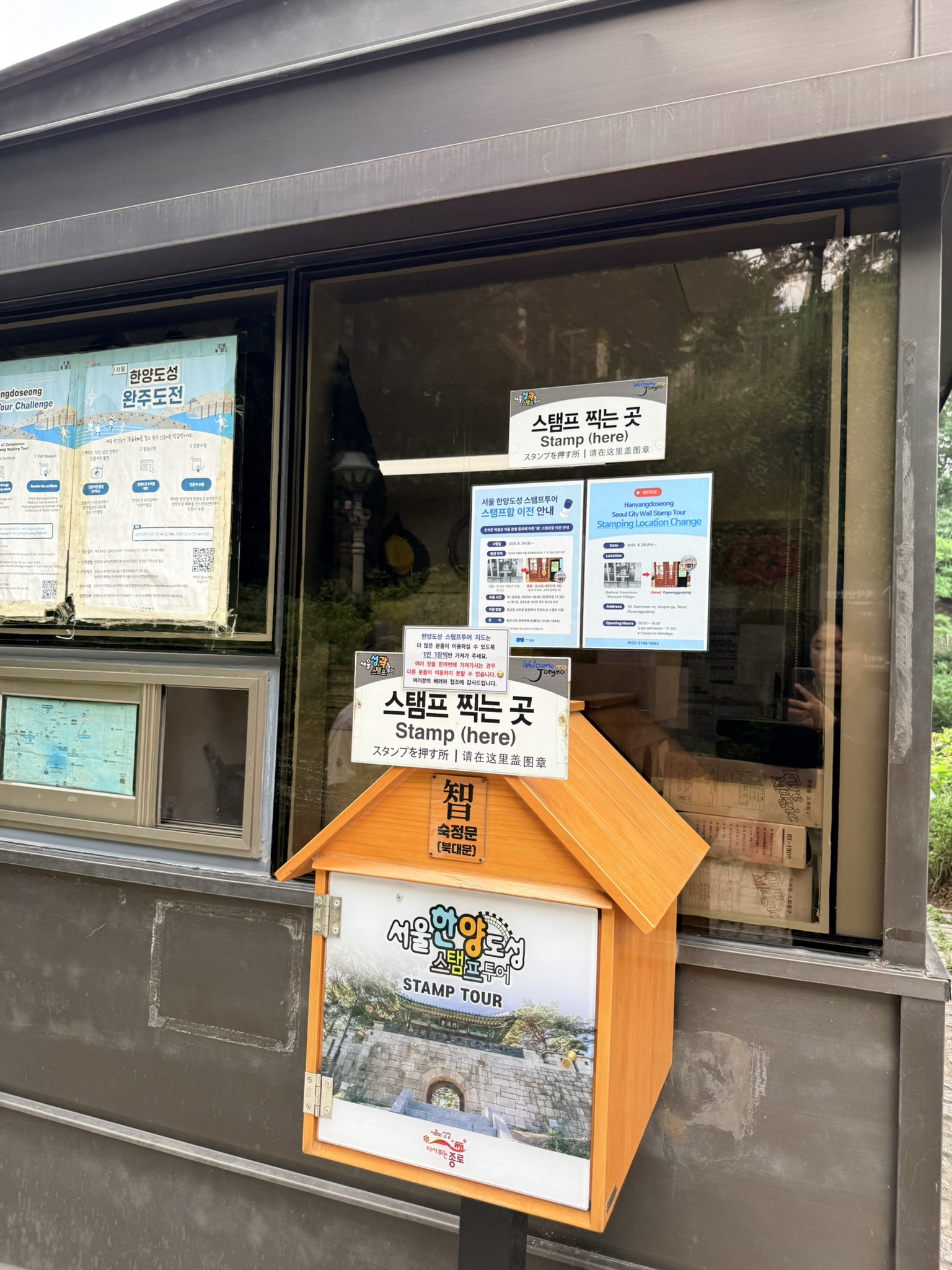 Seoul City Wall Stamp Tour wooden house-shaped stamp box labeled in Korean, English, Japanese, and Chinese mounted outside a visitor kiosk window at Sukjeongmun, Hanyangdoseong tour challenge materials visible through the glass