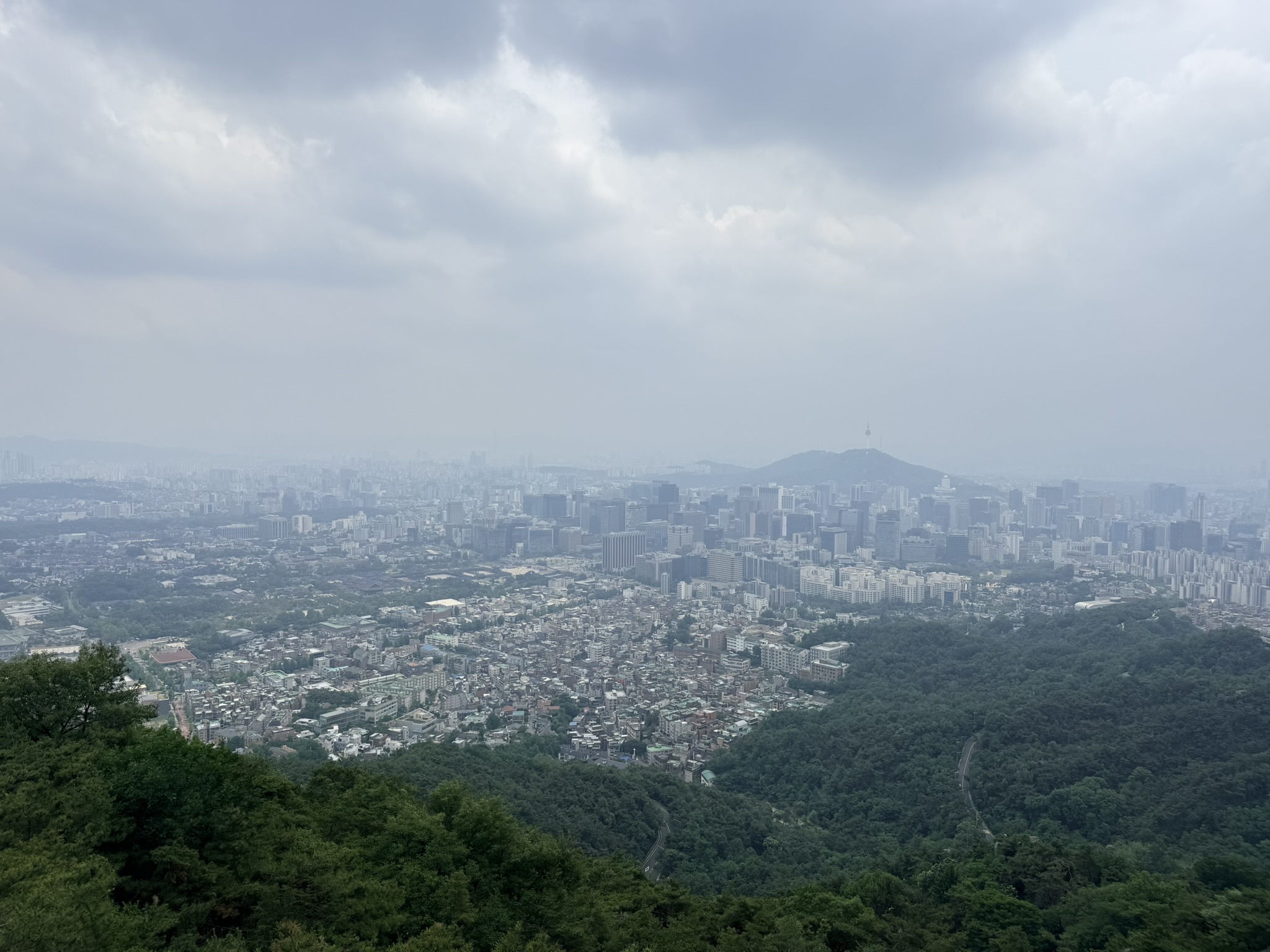 Wide panoramic view of Seoul from the Inwangsan trail under overcast skies, N Seoul Tower silhouette visible on Namsan hill in the mid-distance, green ridge in the foreground