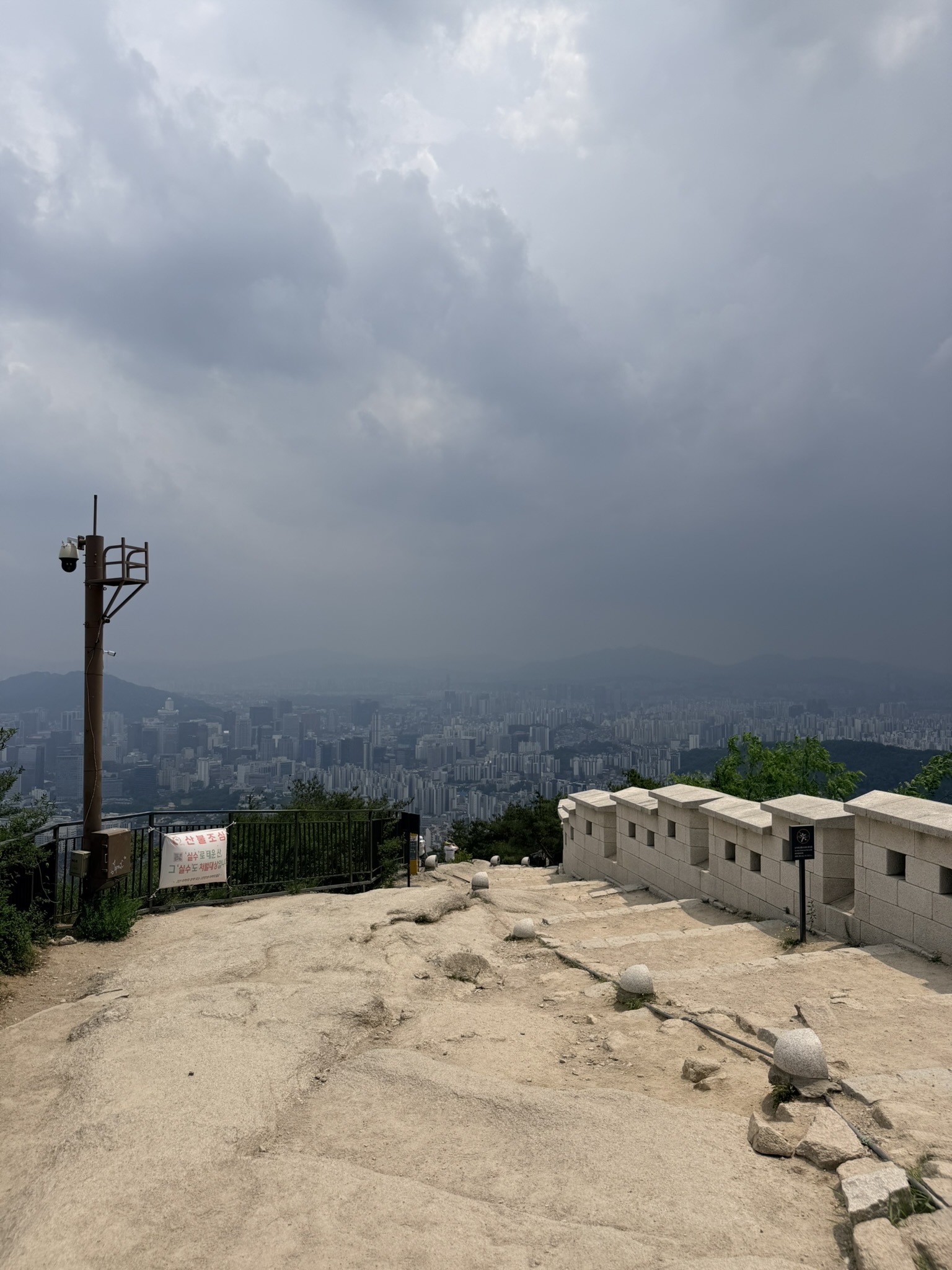 Bare granite summit plateau of Inwangsan at 338.2m with Hanyangdoseong battlements, CCTV pole, forest fire warning banner, and dark storm clouds massing over the Seoul cityscape below