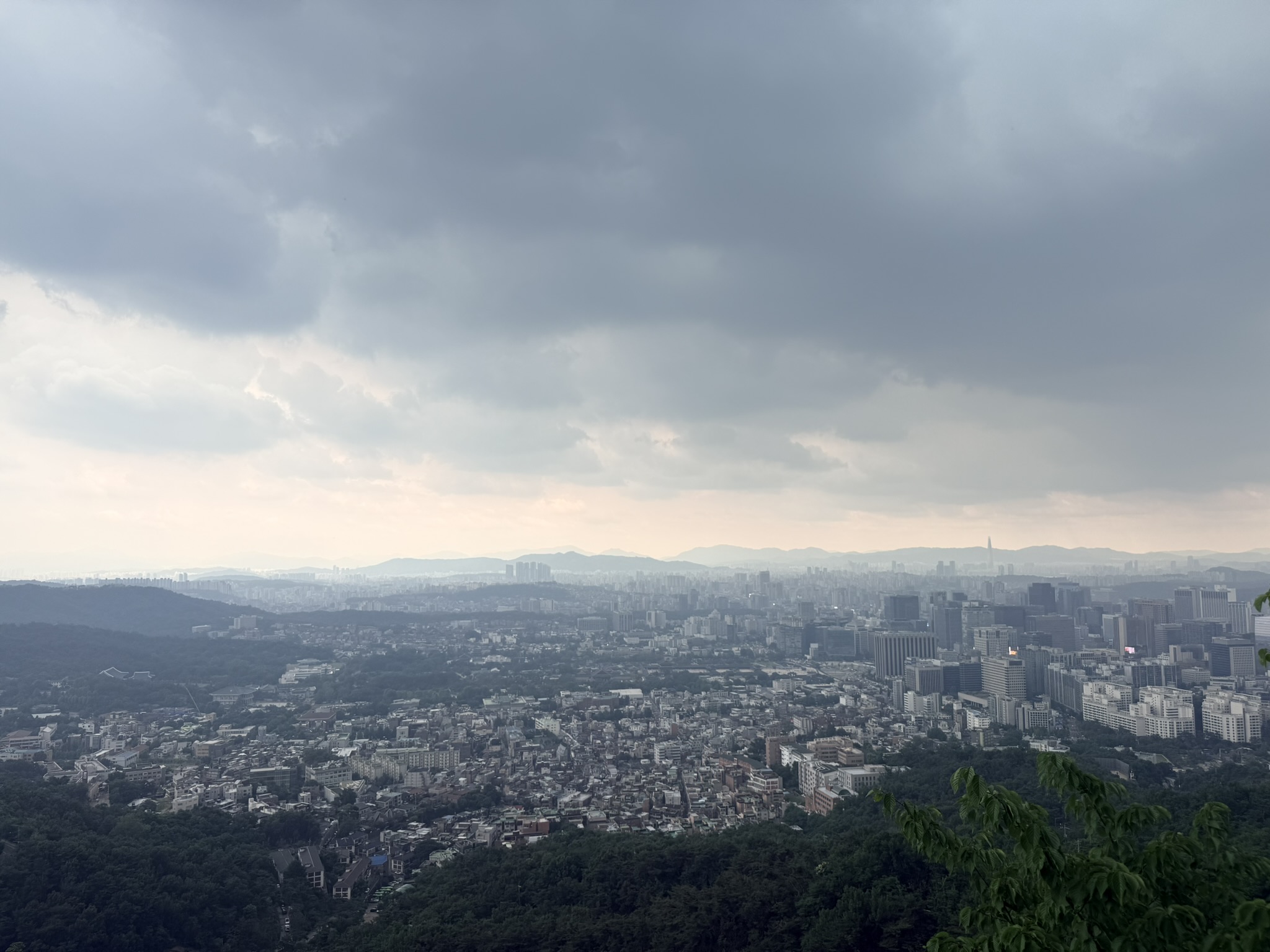 Wide panoramic view from Inwangsan summit showing Seoul spread under pre-storm silvery light, Lotte World Tower silhouette at the far right, dense urban sprawl extending to distant mountains and the Han River basin